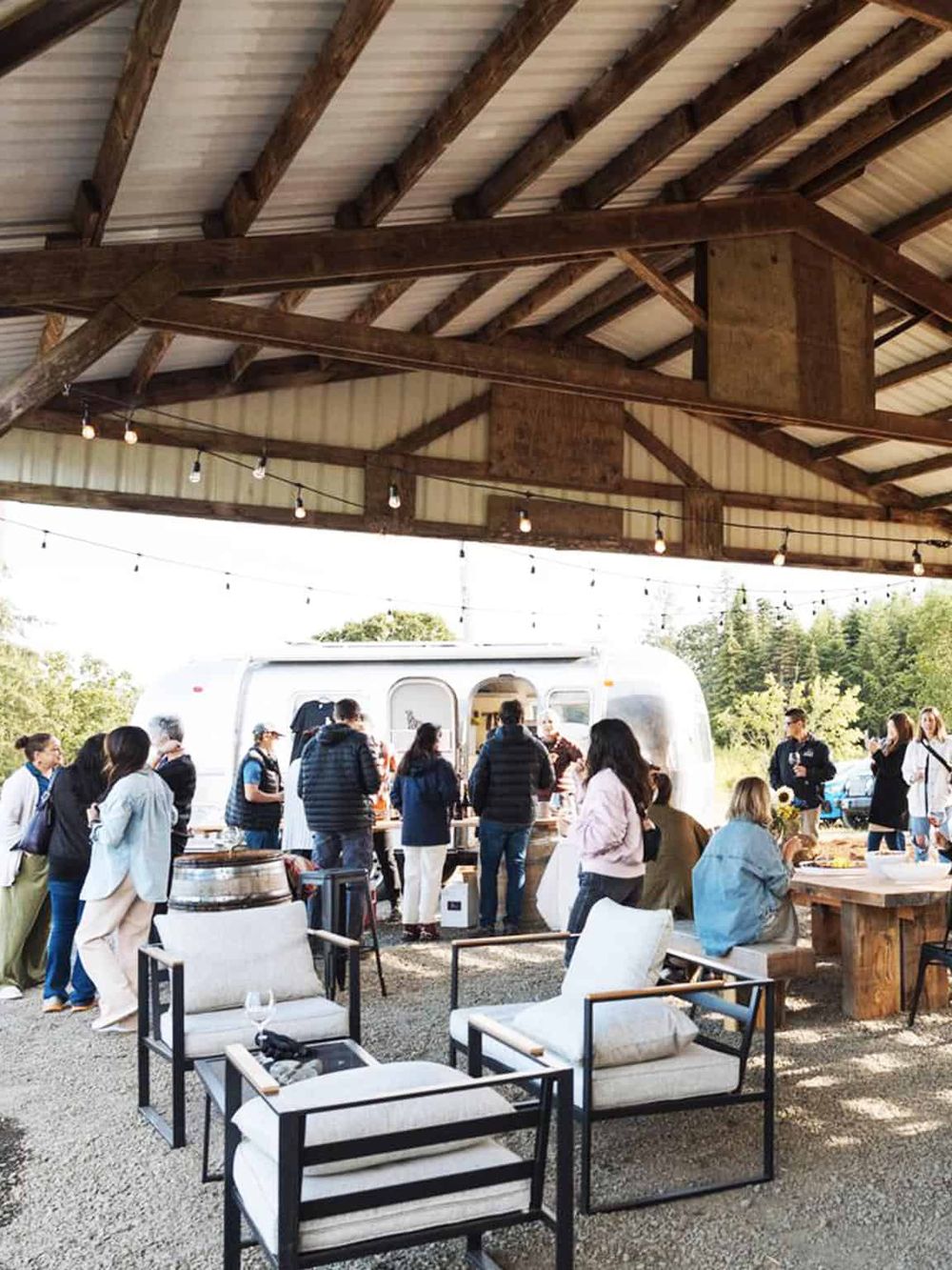 Outdoor event with food trucks and people socializing under a wooden pavilion.