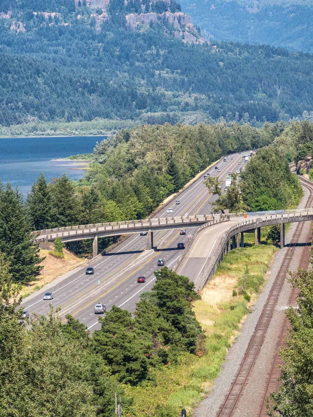 Scenic highway with cars driving alongside a river and lush green forested mountains.