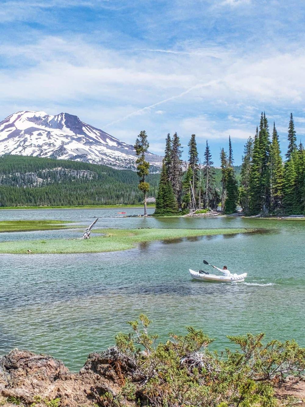 Serene mountain lake with snow-capped peak, pine trees, and kayaker enjoying outdoor adventure.