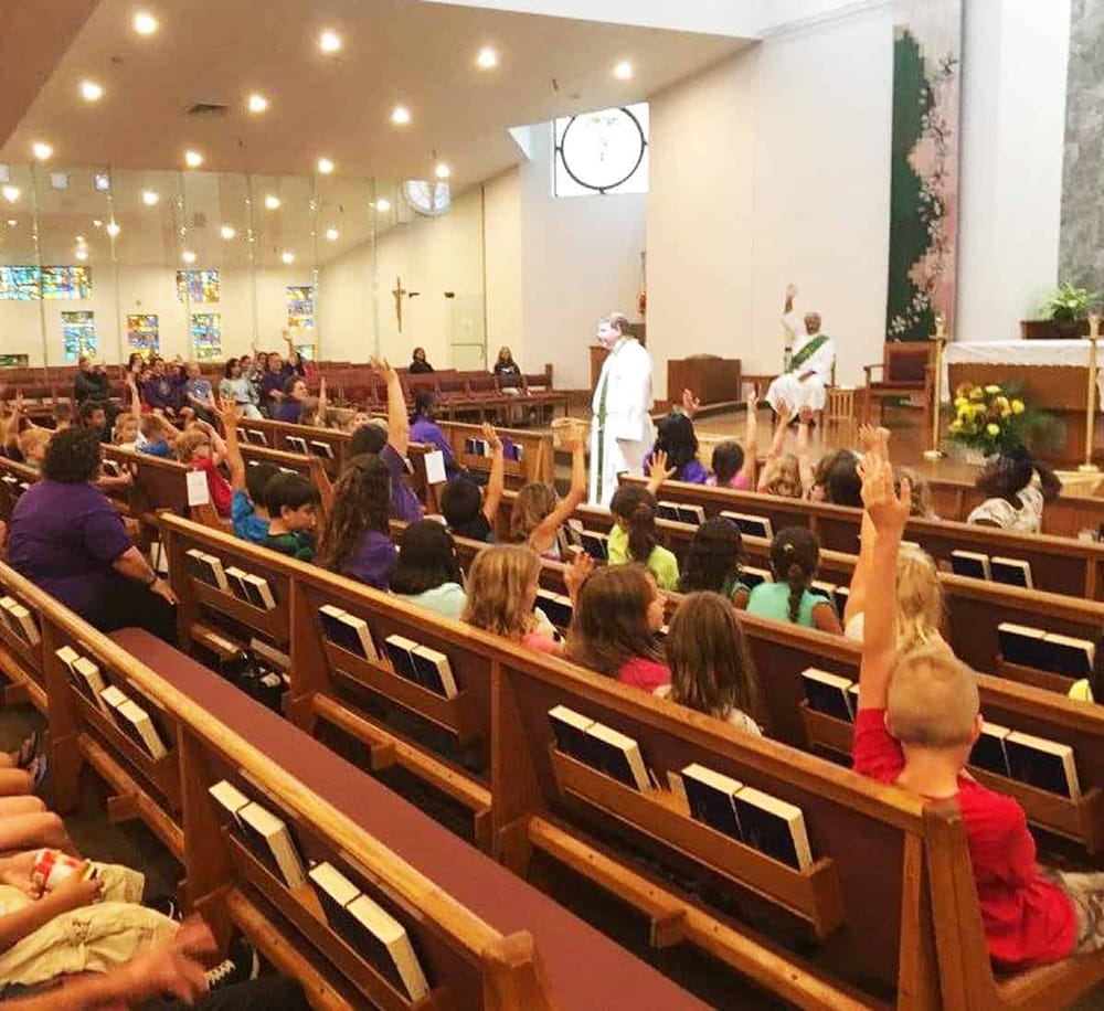 Children raising hands during church service in a modern, well-lit sanctuary with stained glass windows.