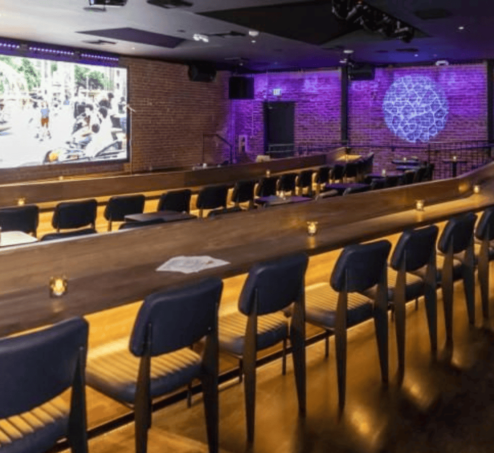A cozy bar and lounge area with ambient lighting, brick walls, and a large screen for live sports or entertainment.