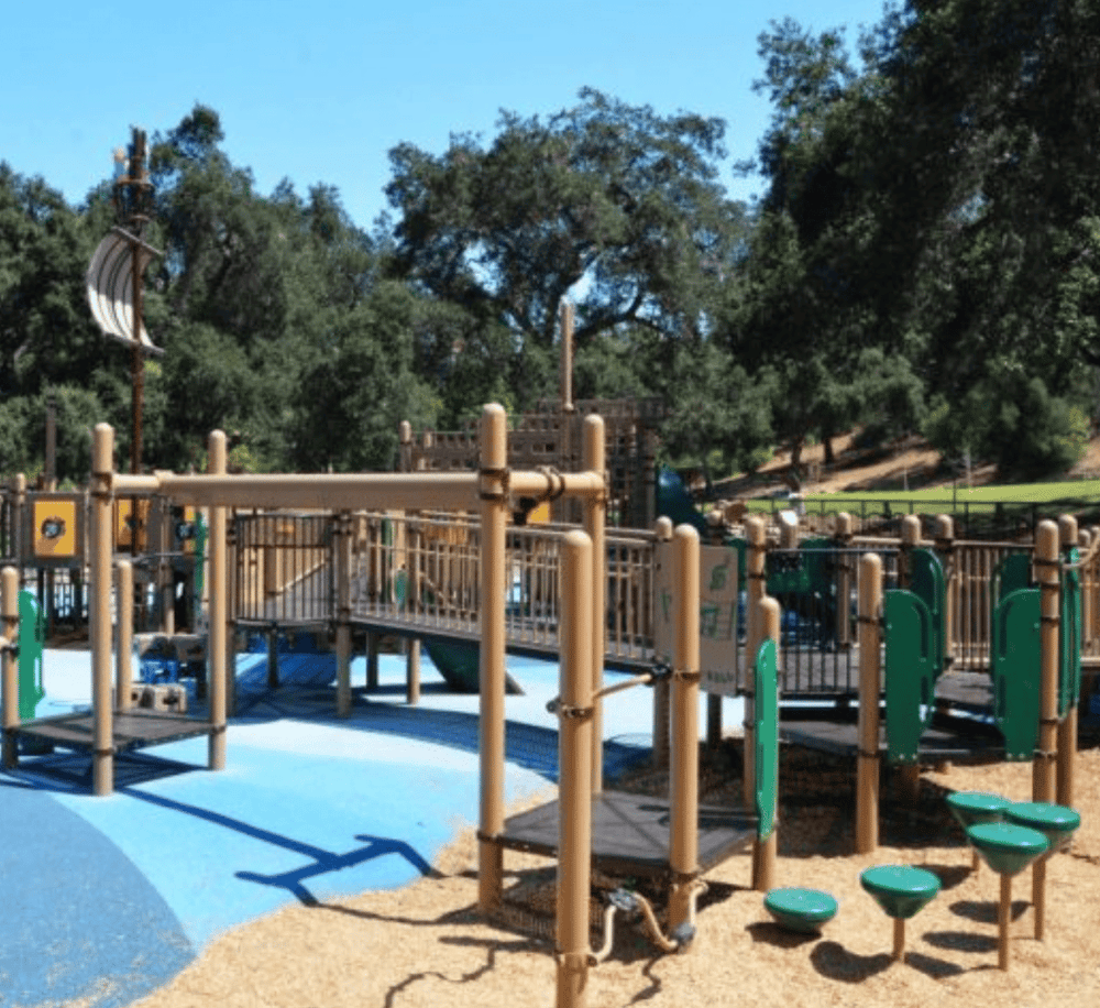 Colorful outdoor playground with wooden structures and rubber safety surface in park setting.