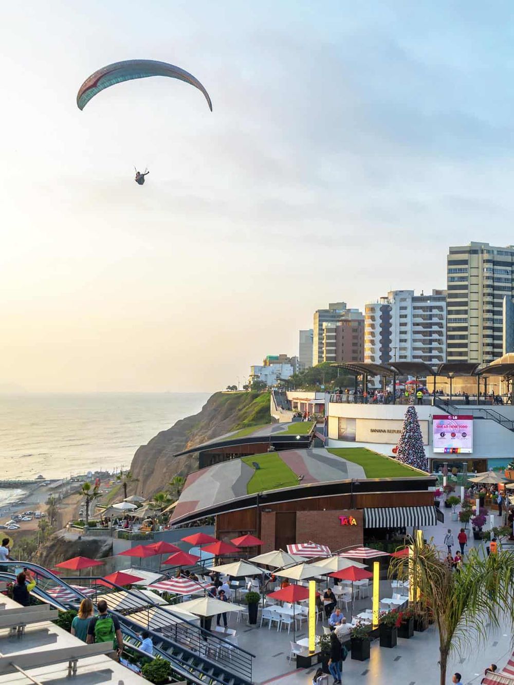 Parasailing over scenic coastal cityscape with modern buildings and vibrant outdoor cafes at sunset.