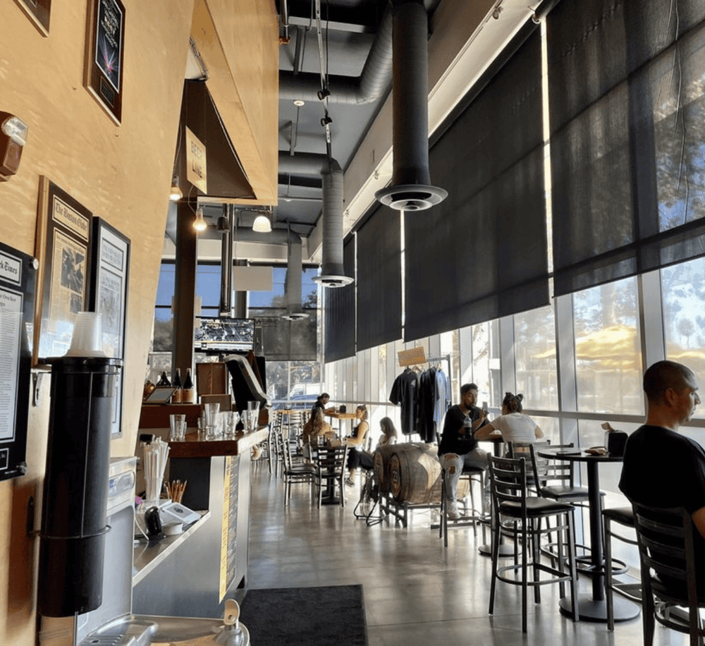 Modern coffee shop interior with large windows, black window shades, and patrons enjoying drinks and conversation.