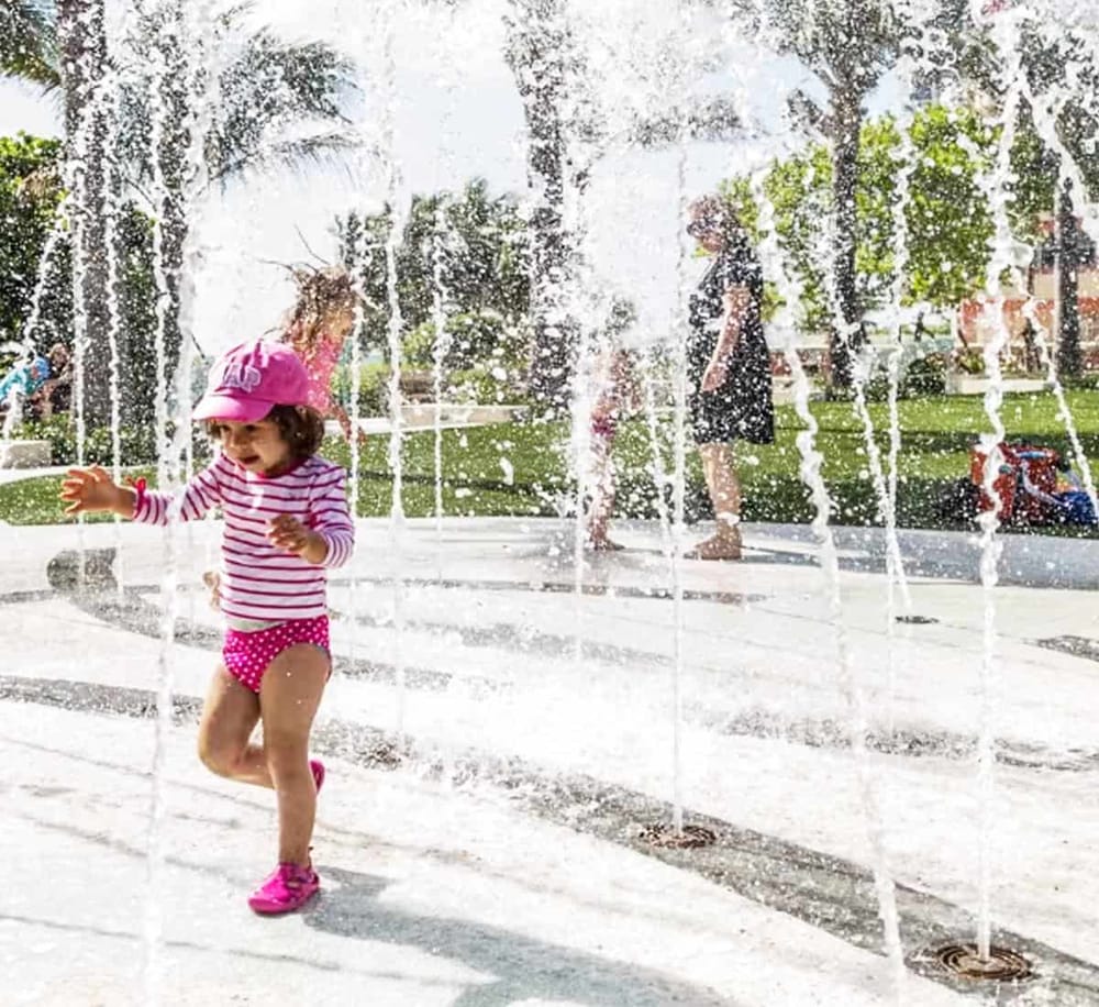 Bright young girl playing in water fountain on a sunny day, family-friendly outdoor activity.