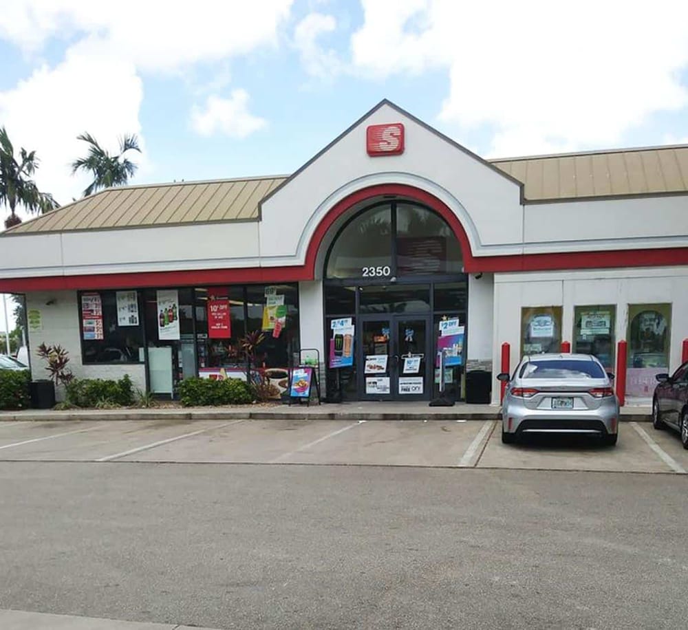 Convenience store with red and white exterior, parking lot, and palm trees in the background.