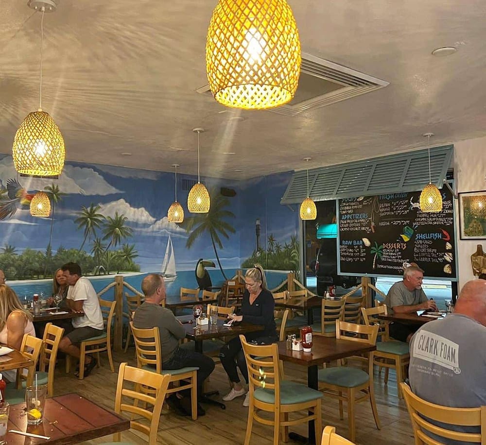 Vibrant tropical-themed restaurant interior with ocean mural, hanging wicker lights, and diverse diners enjoying meals.