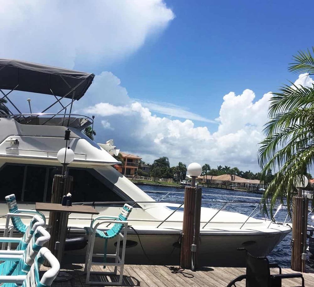 Luxury yacht docked at a scenic waterfront marina with tropical palm trees and residential buildings in the background.