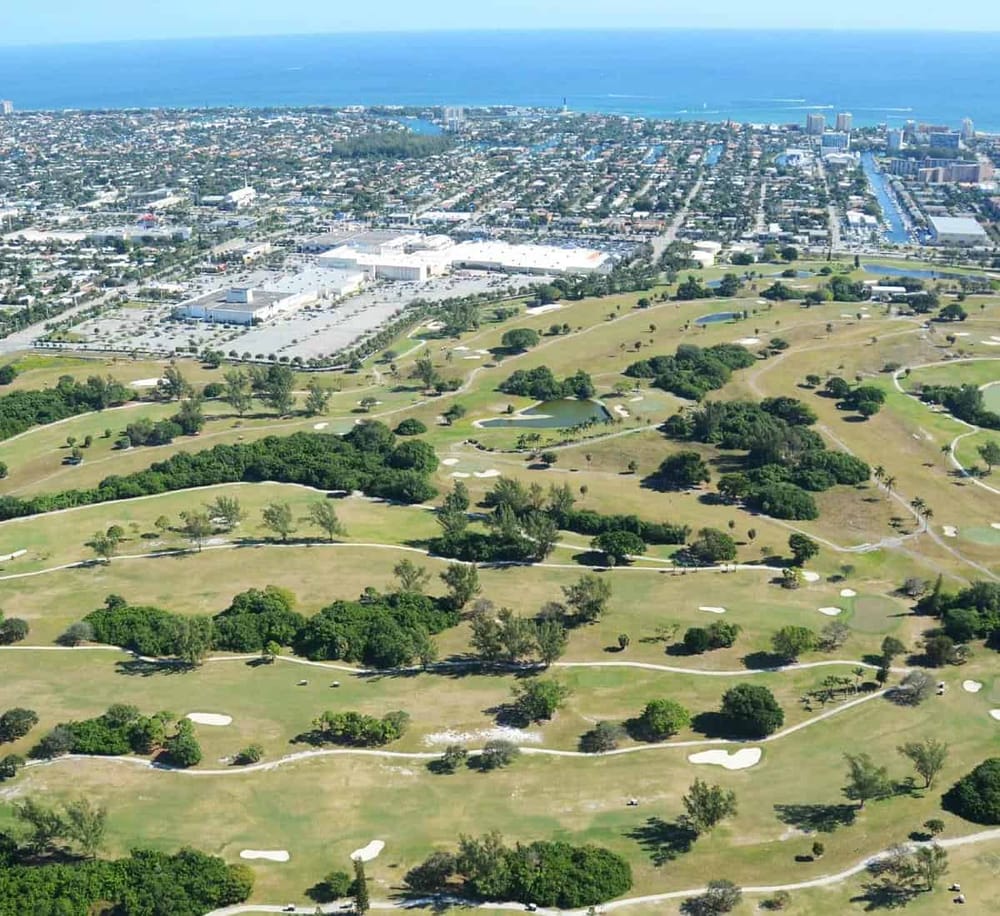 Open-air golf course with lush greens and cityscape background, near the coast.