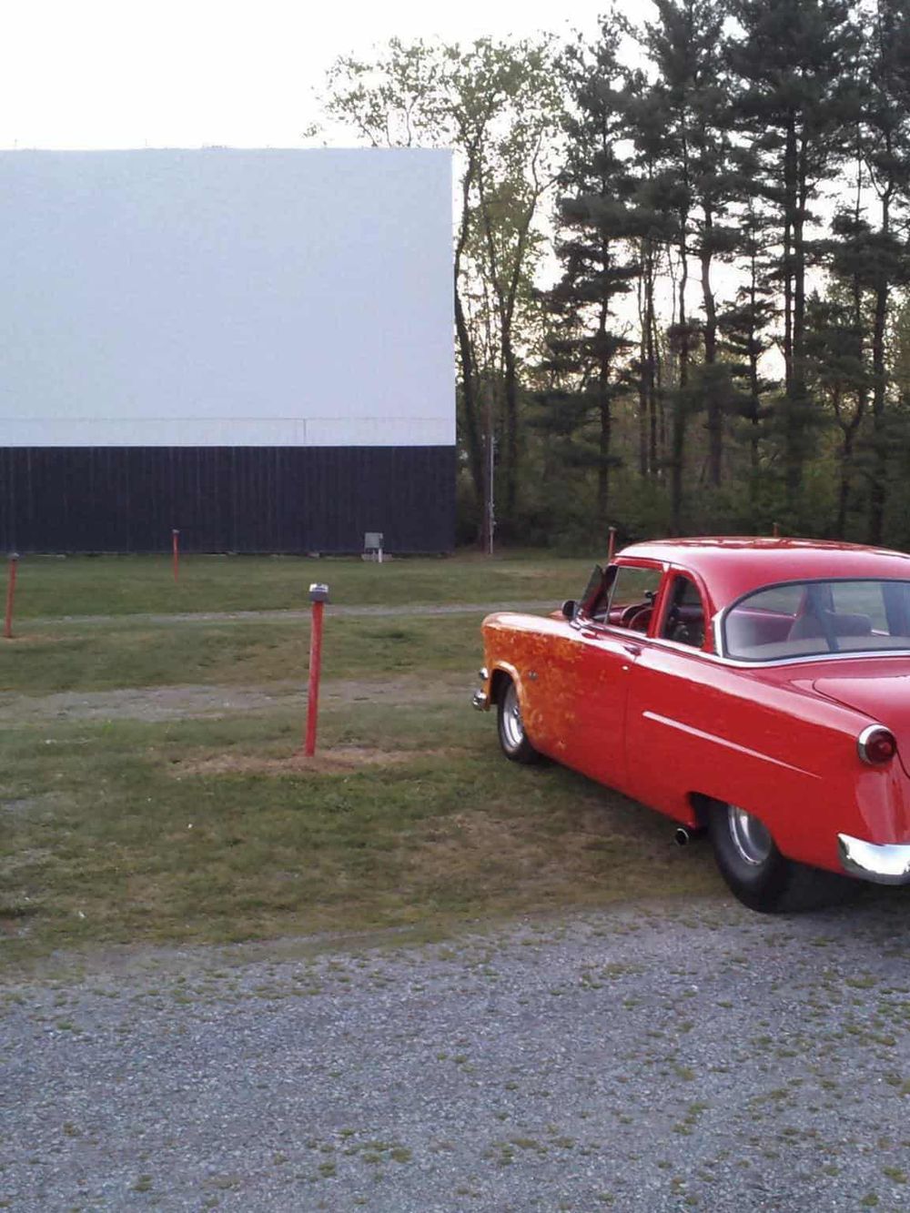 Classic vintage red car parked near an outdoor movie screen in a scenic natural setting.