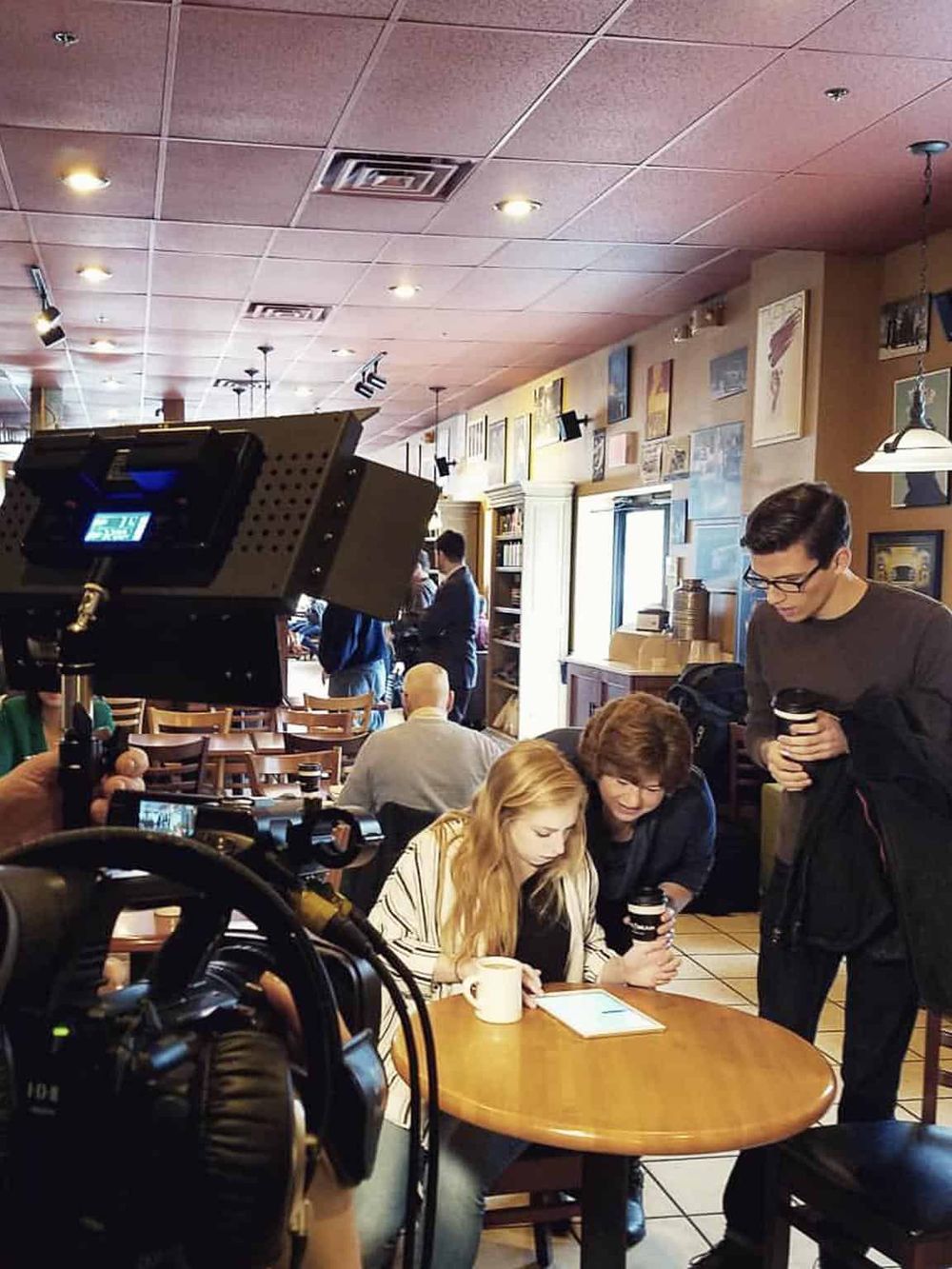 Candid shot of people filming a cozy coffee shop with baristas, art, and customers enjoying beverages.