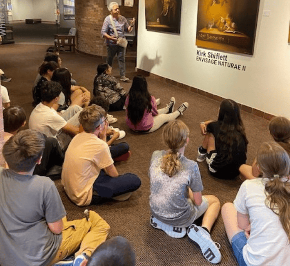 Children sitting on the floor listening to an art gallery tour by a guide.