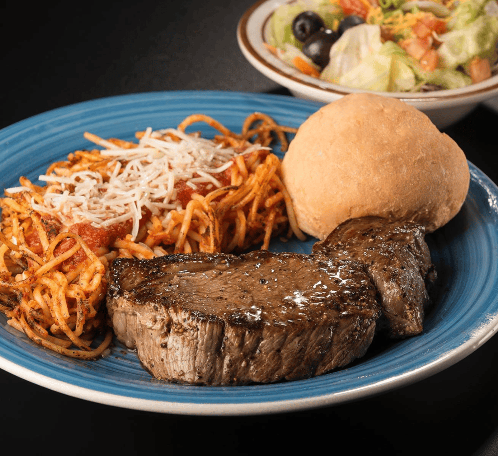 Juicy steak, pasta, bread roll, and salad on blue plate at a restaurant.