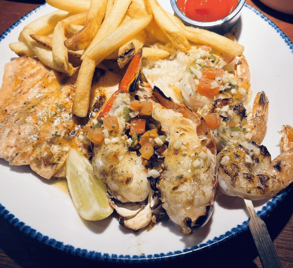 Juicy grilled lobster tails with seasoned toppings, French fries, and lemon wedge on a white plate.