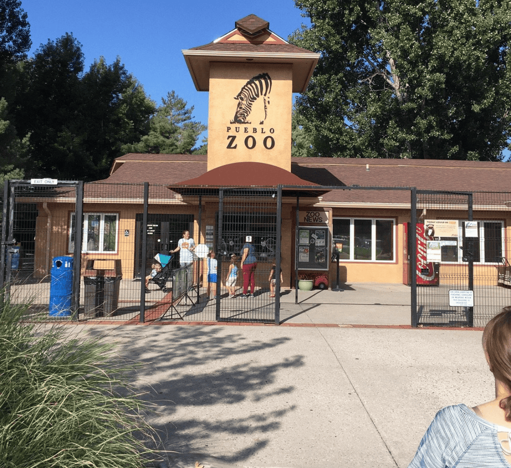 Colorful zoo entrance with children and families, Pueblo Zoo signage, outdoor setting, and gated entry in a scenic park area, ideal for family adventures.