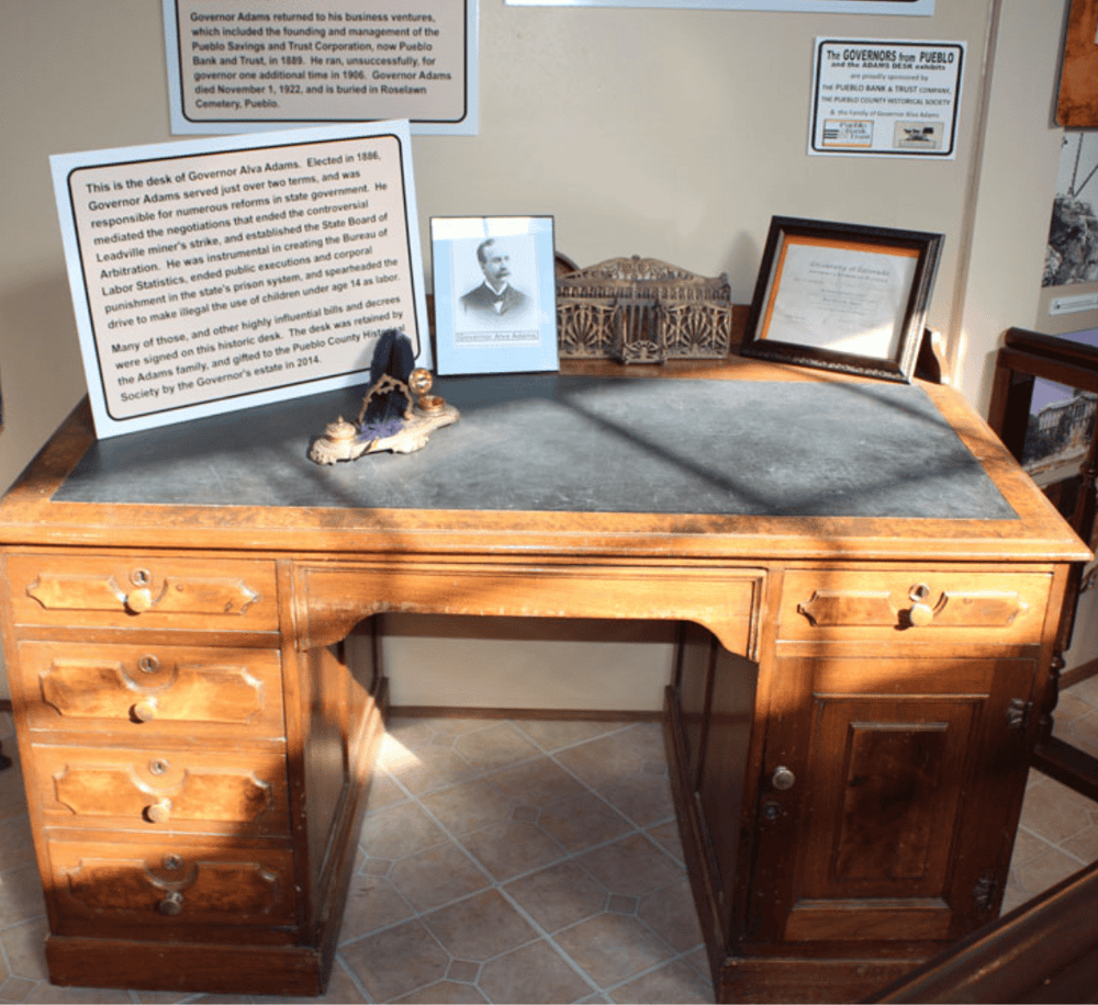 Alto text: Antique wooden desk with historical plaques and framed photographs in a museum setting.