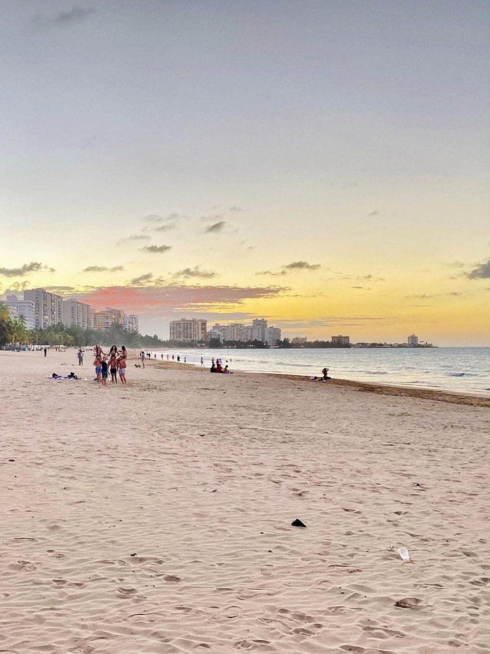 Sunset beach scene with people relaxing and socializing near skyscrapers, showcasing coastal city life and seaside leisure.