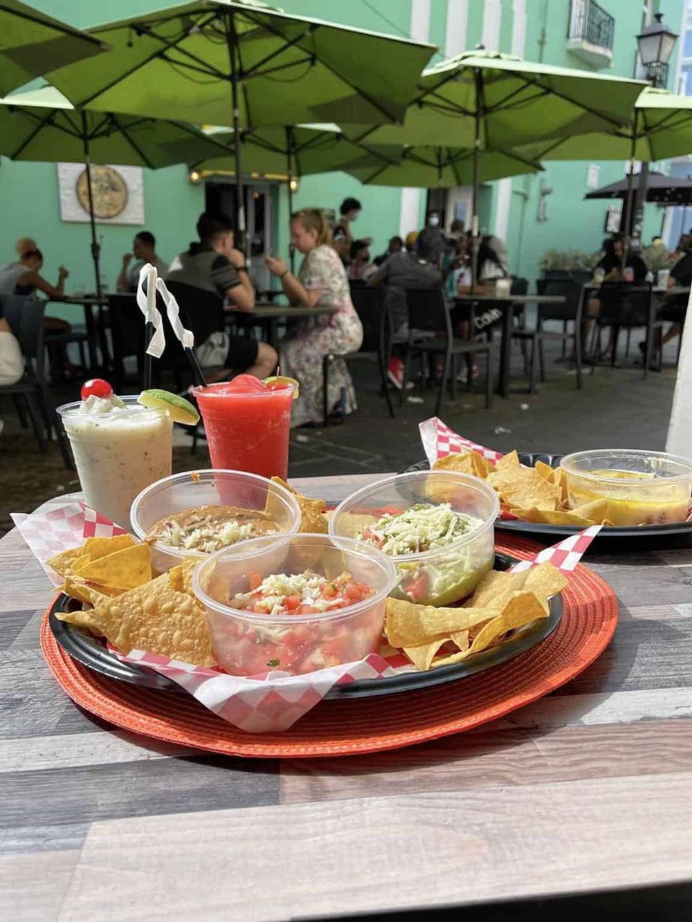 Colorful Mexican food and drinks served on a tray at an outdoor restaurant with green umbrellas in a lively city setting.