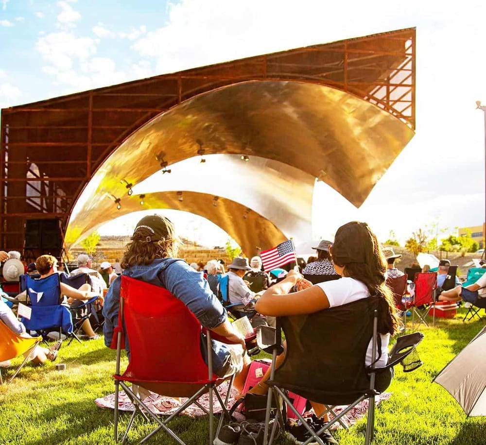 People enjoying an outdoor concert with a modern stage featuring a gold, curved design, on a sunny day.
