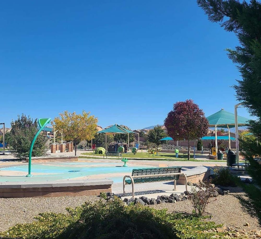 Colorful park with shade umbrellas, benches, and children’s water play area under clear blue sky.