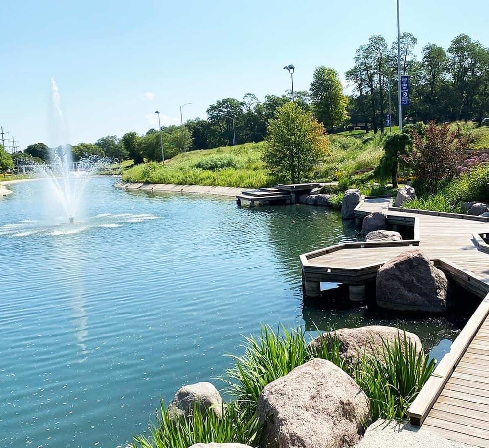 Serene park pond with wooden docks, water fountain, lush greenery, and walking paths, perfect for outdoor relaxation.