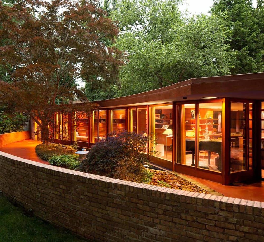 Modern glass house with warm interior lighting amid lush greenery and mature trees.