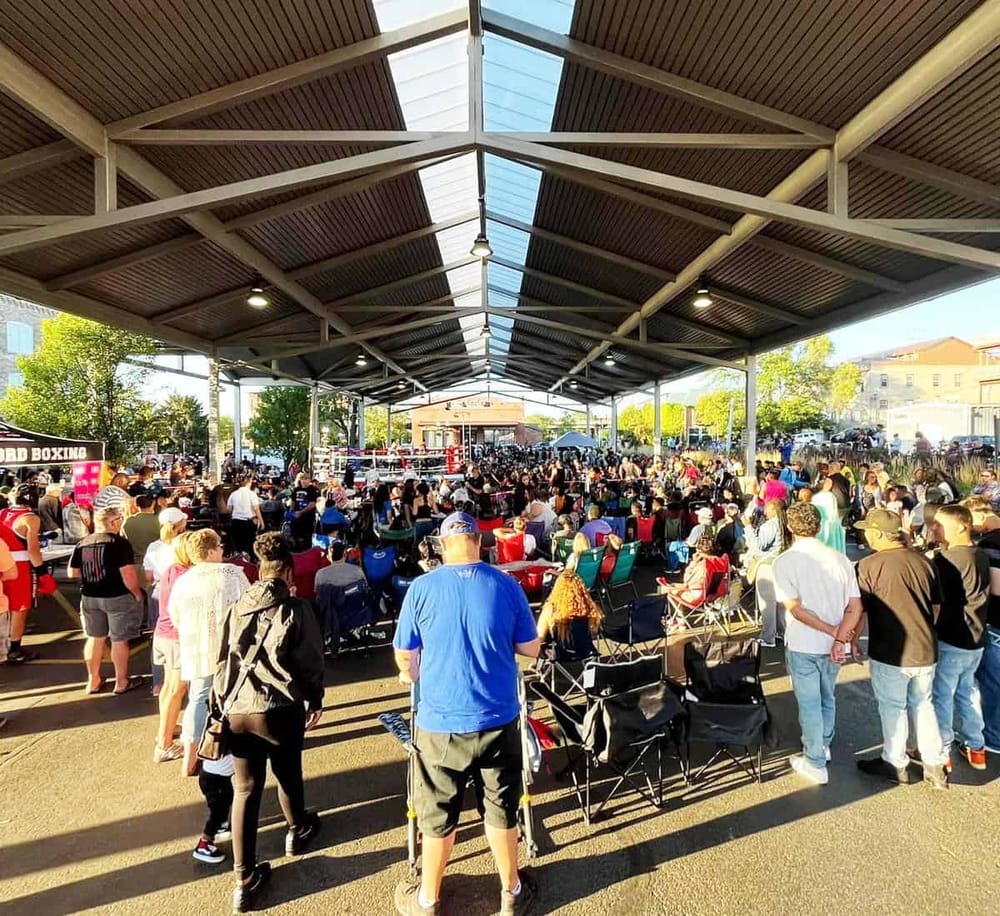 Spectator crowd gathered at an outdoor boxing event under a large covered pavilion.