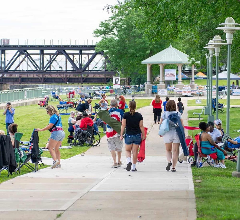 Bright outdoor scene at a community event in a park with walking paths, people gathering, tents, and a bridge in the background.