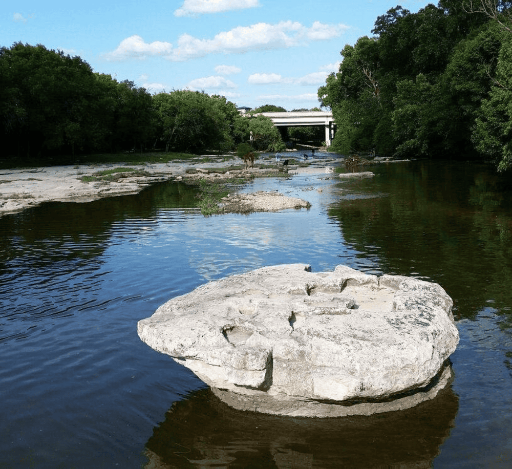 Tranquil river scene with large rock, lush green trees, and a bridge in the background for outdoor adventure.