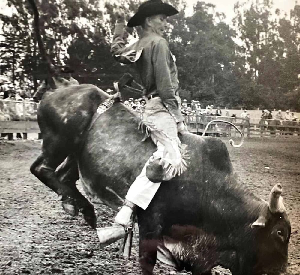 Rodeo cowboy riding a bull at a rodeo event with spectators in the background.