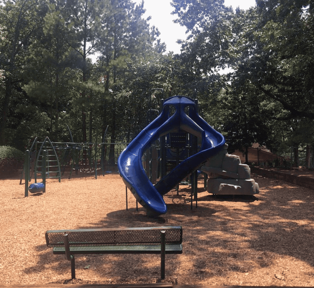 Bright blue playground slide at Quest For Directions park, surrounded by trees and shaded areas.