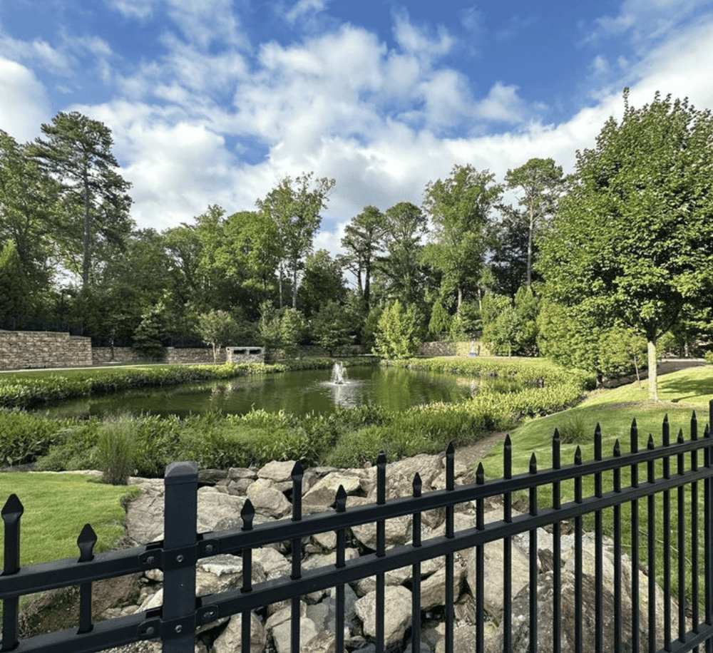 Serene park pond view with fountain, lush greenery, trees, and decorative iron fence, peaceful outdoor setting.