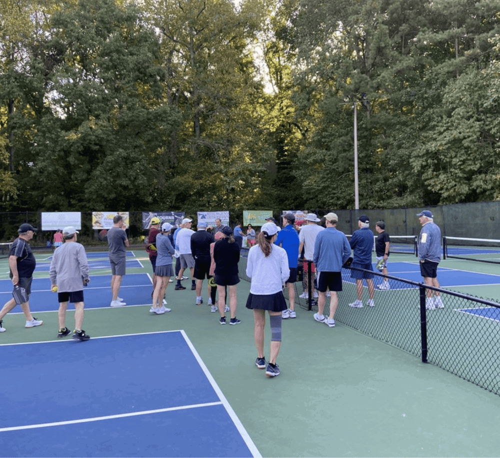 Tennis players gather on courts during a daytime tennis event, surrounded by lush trees.