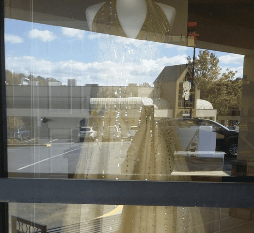 Colorful dress displayed behind a glass window with raindrops, reflecting the parking lot and buildings outside.