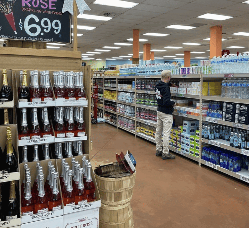 1. Wide shot of grocery store aisle with beverages, water bottles, and shopper browsing products.