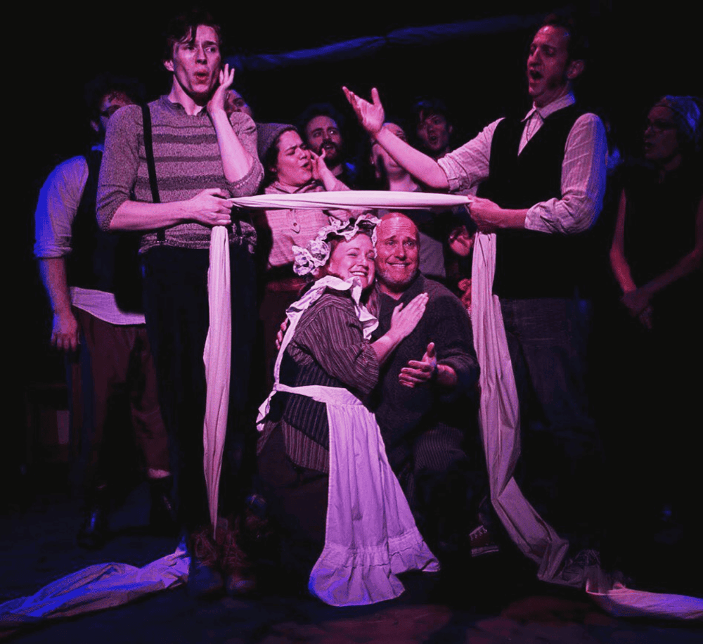 A professional theater performance with actors in period costumes, expressive gestures, and emotional expressions on stage.