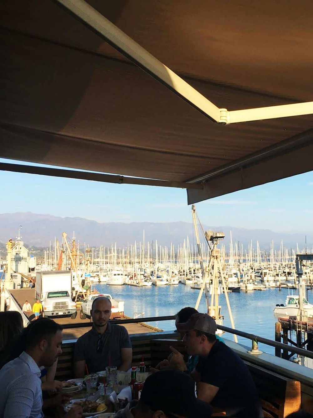 Boaters dining at seaside restaurant with view of marina and mountains in the background.