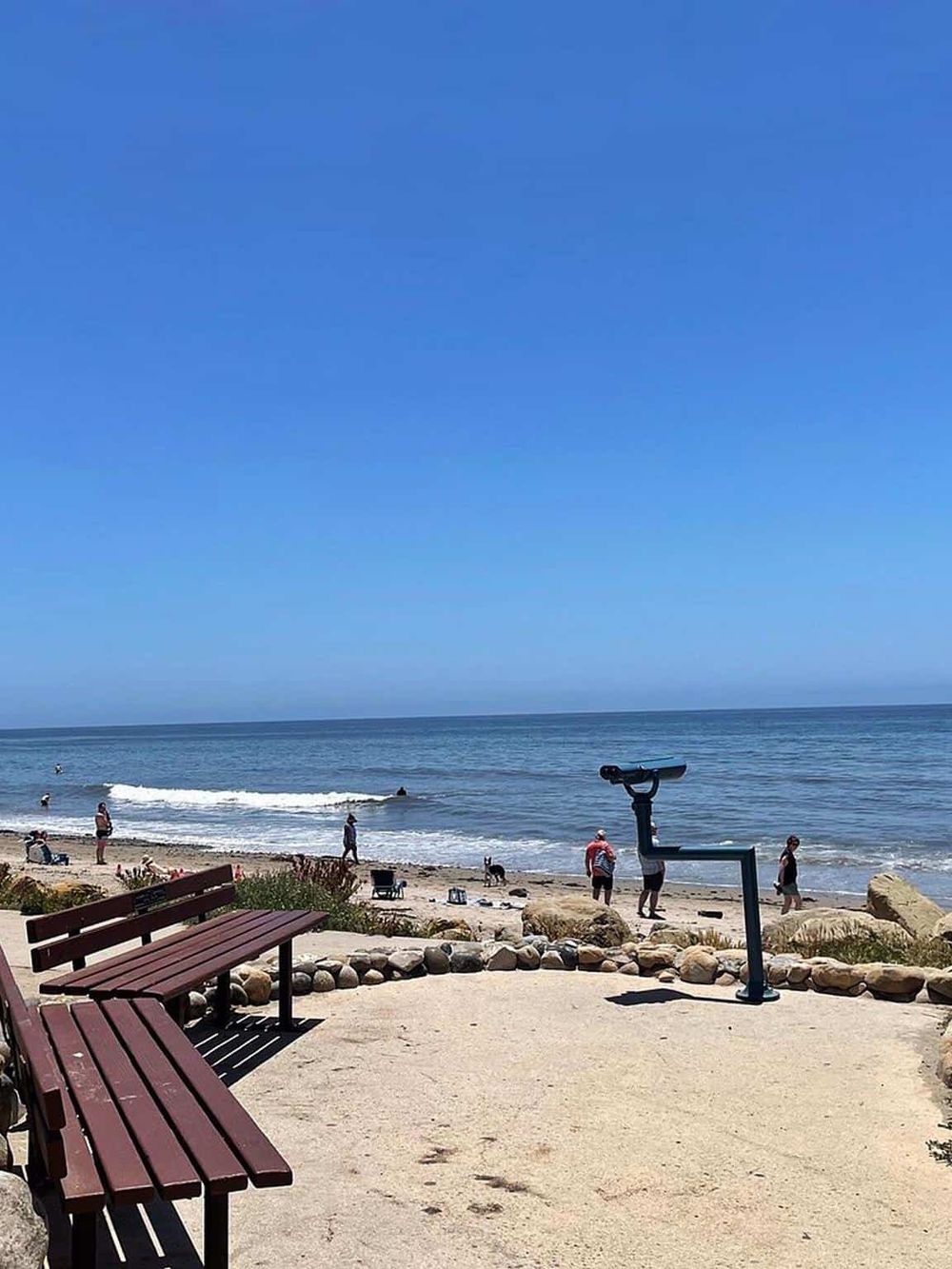Scenic beach with benches, binocular viewer, and people enjoying the ocean view and sunny weather.