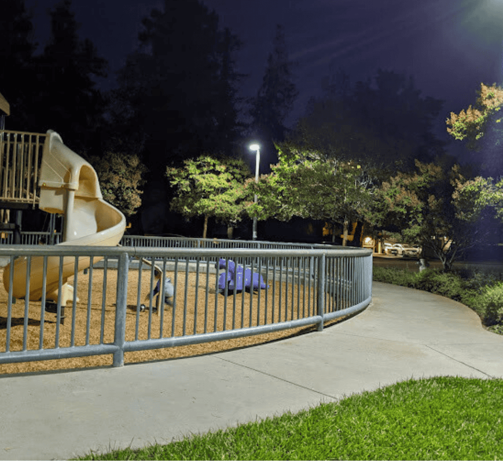Playground slide at night with park pathway and trees in the background.