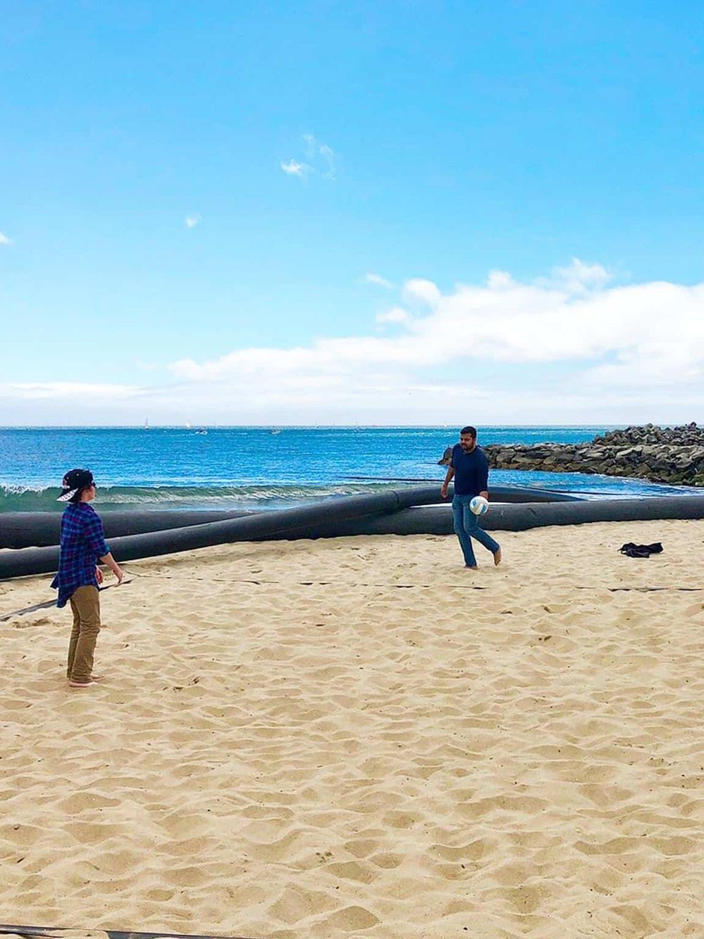 Beach volleyball game on sandy shore with ocean view, two players in casual attire under blue sky.