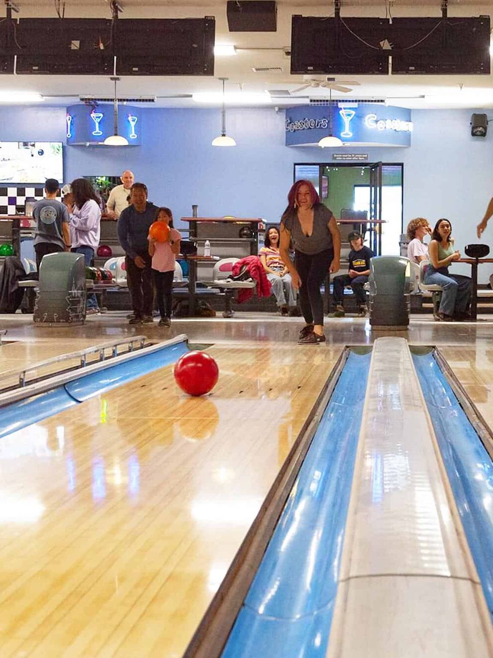 Bright bowling alley with a red ball rolling towards pins, surrounded by smiling players and onlookers.