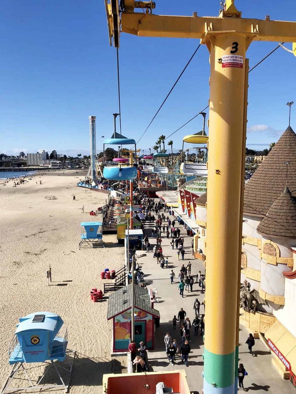 Colorful Ferris wheel above outdoor amusement park on Santa Monica Pier, sandy beach, and visitors enjoying day.