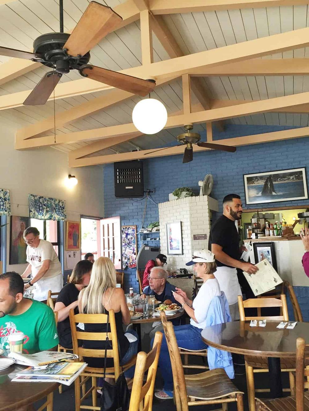 A lively restaurant interior with wood beams, ceiling fans, and patrons dining, reflecting a cozy, welcoming atmosphere.