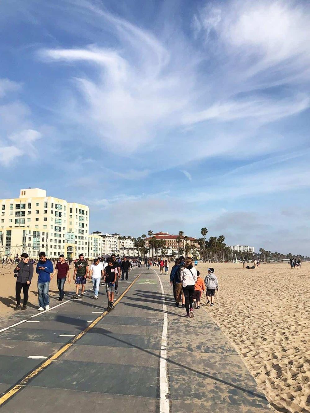 People walking along a beachside promenade with clear skies and modern buildings in the background.