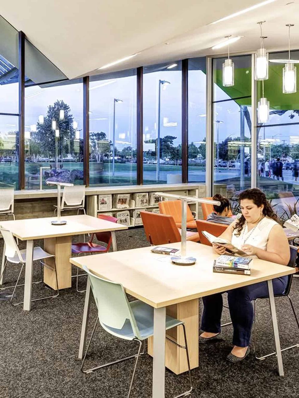 Bright modern library reading area with large windows, soft lighting, and people reading in a quiet space.