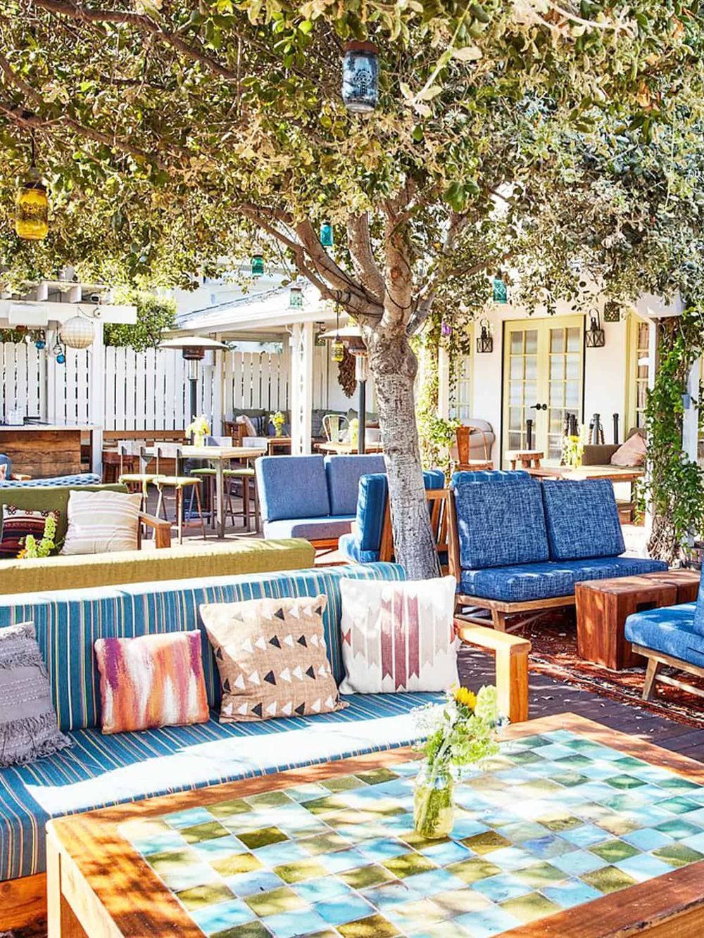 Colorful outdoor patio with string lights, cozy seating, and lush tree for relaxing ambiance.