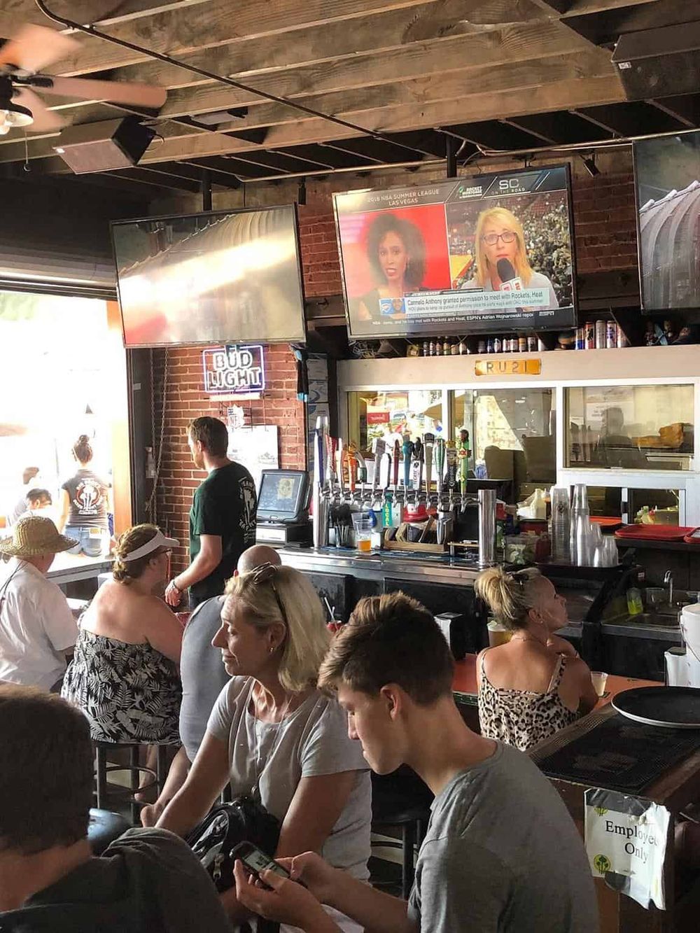 A vibrant bar with multiple TVs, guests watching sports, and a lively atmosphere with wooden decor.