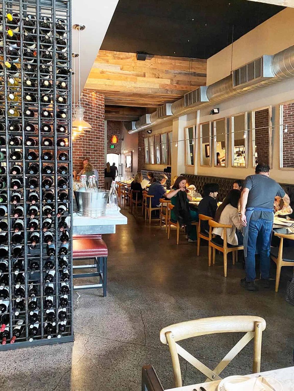 Cozy restaurant interior with wine racks, brick walls, and patrons dining at wooden tables.