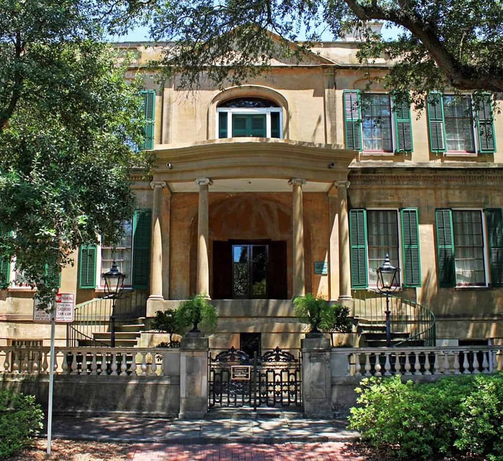 Charming historic building with green shutters and elegant entrance, situated in a lush tree-filled setting.