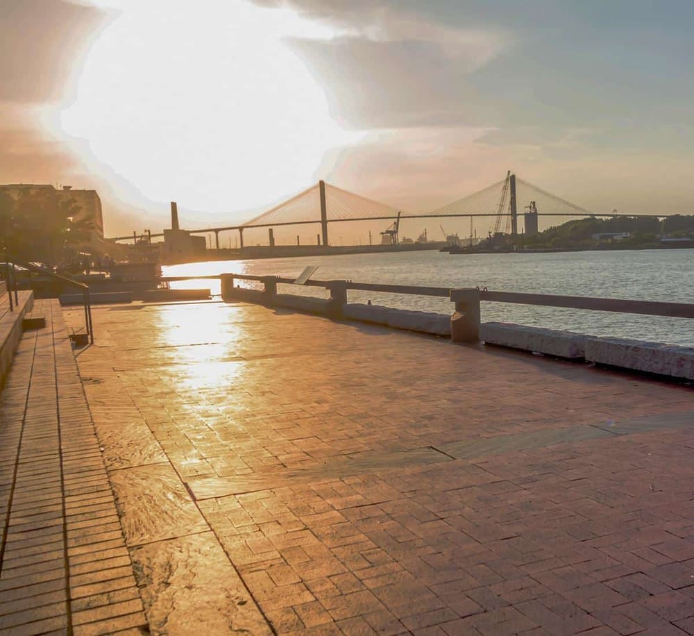 Bright sunset over waterfront promenade with cityscape and bridge in the background.